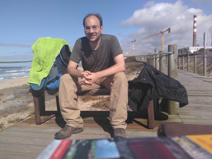 Ein Wanderer sitzt am Rande eines Holzsteges an einem Strand auf einer Bank und schaut in die Kamera, während er die Hände gefaltet hat. Eine Jacke, ein Rucksack und ein Handtuch liegen neben ihm. Im Vordergrund ist verschwommen ein Buch erkennbar, das anscheinend die Kamera stützt. Es ist Sonnenschein, der Wanderer sieht verschwitzt aus, schaut aber zufrieden.