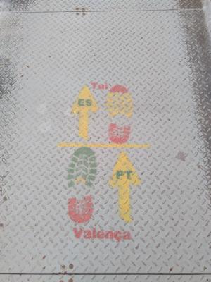 Bodenplatten aus Eisen, vermutlich auf einer Brücke. Aufgesprüht sind zwei Fußabdrücke übereinander, die durch eine waagerechte Linie getrennt sind. Links neben dem oberen Fußabdruck ist ein Pfeil, in dem “ES” steht. Neben dem unteren ist ein Pfeil in dem “PT” steht. Beide zeigen vorwärts. Unter dem Ganzen steht “Valenca”, über dem Ganzen steht “Tui”. Die obere Hälfte ist in den spanischen Farben gesprüht, die untere Hälfte in den portugiesischen.