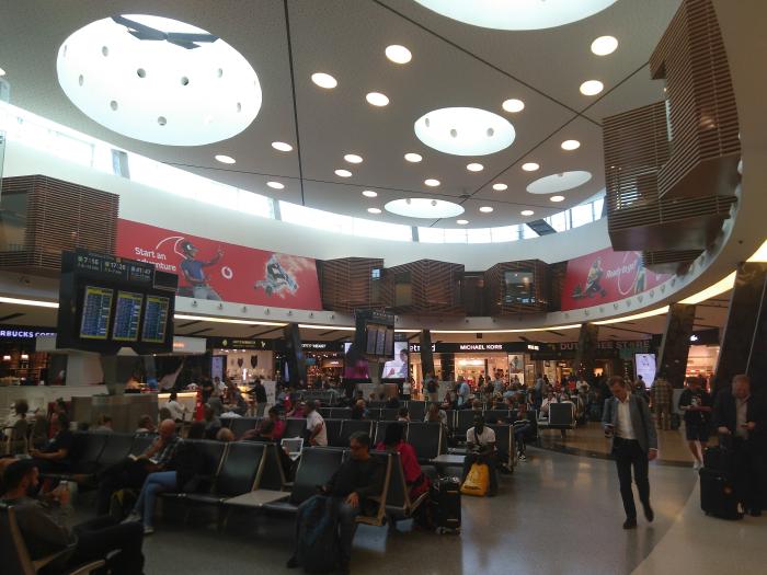 Eine Abflughalle in einem Flughafen mit vielen Menschen. In der Mitte Sitzgelegenheiten, am Rande Geschäfte und Monitore mit Abflugtableaus.