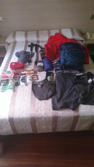Eine Wanderausrüstung auf einem Bett ausgebreitet. Sichtbar sind u.a. Rucksack, Regenjacke, Hose, Wanderstöcke, Erste-Hilfe-Set, Hygieneutensilien und Kleinkram.