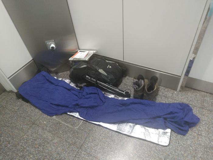 Ein Schlafsack in einer Ecke auf einer dünnen Isomatte aus Alu auf einem Fliesenboden. Daneben ein Rucksack und Wanderschuhe. Auf dem Rucksack liegen Zettel und ein Handy, das an einer Steckdose in der Wand hängt.