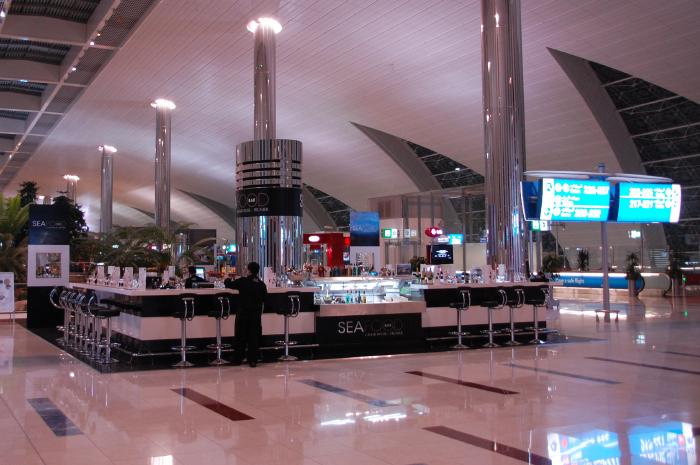 Das Bild zeigt eine moderne, elegante Bar in einem Flughafen. Die Bar ist mit einer weißen und schwarzen Theke, hohen Barhockern und einer Glasvitrine ausgestattet, in der Speisen und Getränke präsentiert werden. Ein Barkeeper ist zu sehen. Die Umgebung wirkt hell und modern, mit hohen Säulen und einer auffälligen Decke. Im Hintergrund befinden sich Abflugtafeln und Grünpflanzen. Das Gesamtbild ist geprägt von Reinlichkeit, Luxus und Funktionalität, typisch für eine Flughafen-Lounge.