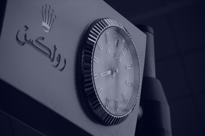 Das Bild zeigt eine Rolex-Uhr, die an einer Wand befestigt ist. Die Uhr ist silberfarben und hat ein gewelltes Zifferblatt. Neben der Uhr befindet sich das Rolex-Logo und arabische Schrift. Das Bild ist in Sepia gehalten.