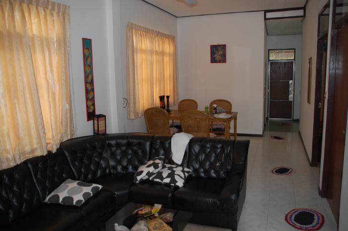 Das Bild zeigt ein Wohnzimmer mit einer schwarzen Ledercouch im Vordergrund. Auf dem Sofa liegen einige Kissen. Im Hintergrund befindet sich ein Esstisch mit Stühlen und ein Flur mit Türen. Die Wände sind hell gestrichen, und an den Fenstern hängen beigefarbene Vorhänge. An den Wänden hängen auch einige Bilder. Der Boden ist gefliest. Der Gesamteindruck ist der eines einfachen, aber sauberen und ordentlichen Wohnzimmers.