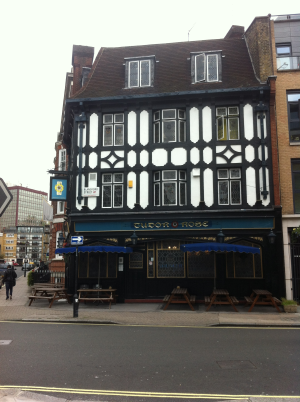Das Bild zeigt einen Pub namens Tudor Rose. Es ist ein Fachwerkhaus mit schwarz-weißer Fachwerkkonstruktion. Vor dem Pub stehen einige Holztische und -bänke, auf denen man draußen sitzen kann. Die Straße vor dem Pub ist mit gelben Linien markiert. Im Hintergrund sind weitere Gebäude zu sehen.