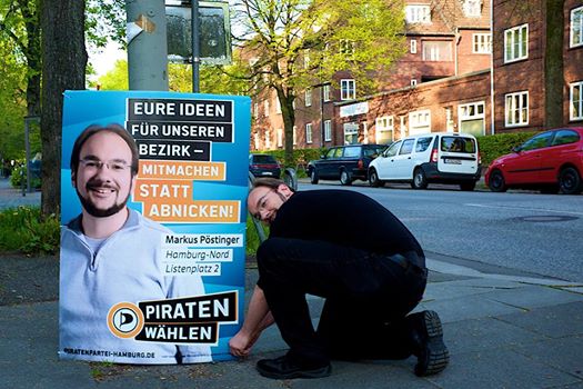 Markus Pöstinger kniet lächelnd an einem Laternenmast neben einem Wahlplakat der Piratenpartei mit seinem Porträt und dem Slogan „Eure Ideen für unseren Bezirk – Mitmachen statt Abnicken!“; im Hintergrund eine Straße mit parkenden Autos und Backsteinhäusern.