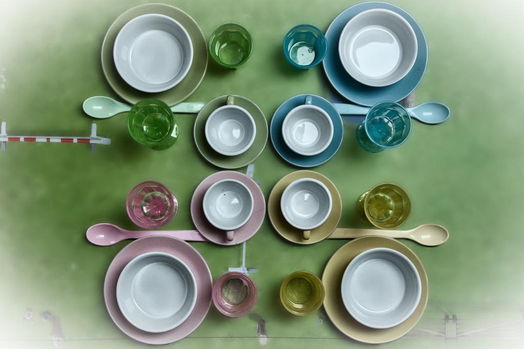 Eine Aufnahme von Kindergeschirr auf einem grünen Spieltisch. Es sind insgesamt vier Sets in gelb, grün, rosa und blau, die quadratisch angeordnet sind. Auf einem großen (in Kindergröße) Teller steht eine kleinere Suppenschüssel. Oberhalb der beiden Teile liegt jeweils quer ein Löffel Richtung außen. Oberhalb des Löffels ist ein Trinkbecher. Innen neben dem Teller steht ein Trinkglas. Innen oberhalb neben dem Teller steht eine Untertasse mit Tässchen. Alles ist möglichst symmetrisch angeordnet, aber die Geschirrteile sind untereinander ganz leicht windschief, insbesondere die Tässchen.