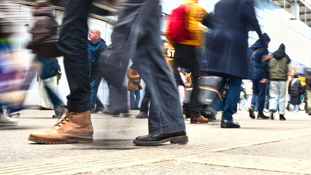 Ein von Bodenhöhe fotografierter und mit vielen Menschen begangener Fußweg, anscheinend an einem Bahnhof oder Flughafen, da einige Koffer sichtbar sind. Die Menschen eilen links und rechts aus dem Bild, die Beine und Körper sind teils verschwommen durch die Bewegung.
