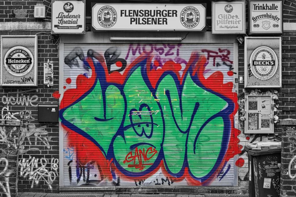 Das Bild zeigt ein Rolltor, das mit Graffiti in Grün, Rot und Blau besprüht ist, welches sich vor einer mit Werbeplakaten und zusätzlichen Graffiti versehenen Backsteinmauer befindet.
Das Bild zeigt ein graues Rolltor aus Metall, an dem ein großes, auffälliges Graffiti in verschiedenen Farben angebracht ist. Das Graffiti ist hauptsächlich in Grün, Rot und Blau gehalten und weist verschiedene, miteinander verbundene Buchstaben- und Formkombinationen auf. Im unteren Bereich des Rolltors ist deutlich das Wort „GANG“ zu lesen. Der Stil wirkt wild und dynamisch, mit starken Kontrasten zwischen den Farben. Die Oberflächenstruktur des Rolltors wird durch das Graffiti nicht verdeckt.
Das Rolltor ist in eine Backsteinmauer eingebettet, an der mehrere Werbeschilder für verschiedene Biersorten angebracht sind. Diese Schilder sind in schwarz-weiß gehalten und stehen im Kontrast zu den bunten Graffiti. Die Schilder weisen verschiedene Schriftarten und Logos diverser Marken auf.