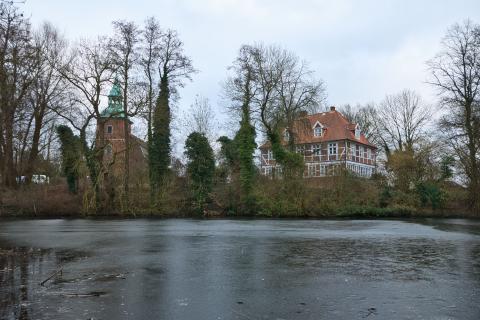 Das Bild zeigt einen zugefrorenen Teich, hinter dem sich ein Backsteingebäude und ein Kirchturm befinden. Die Gebäude liegen auf einer kleinen Anhöhe, die von Bäumen umgeben ist. Die Bäume sind kahl und es ist Winter. Der Himmel ist bedeckt und grau. Das Bild wirkt ruhig und etwas melancholisch.