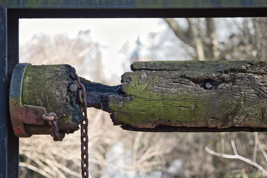 Das Bild zeigt einen alten, verwitterten Holzbalken, der an einer Metallkonstruktion befestigt ist. Der Balken ist an einer Stelle gebrochen und mit einer rostigen Kette verbunden, wie bei einem alten Brunnen. Im Hintergrund ist eine unscharf dargestellte Landschaft mit Bäumen und einem Gebäude zu sehen.