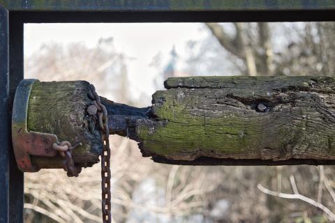 Das Bild zeigt einen alten, verwitterten Holzbalken, der an einer Metallkonstruktion befestigt ist. Der Balken ist an einer Stelle gebrochen und mit einer rostigen Kette verbunden, wie bei einem alten Brunnen. Im Hintergrund ist eine unscharf dargestellte Landschaft mit Bäumen und einem Gebäude zu sehen.