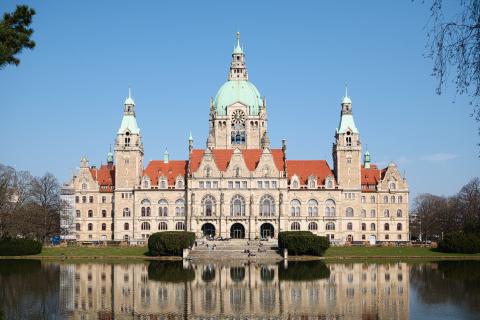 Das Bild zeigt das Rathaus von Hannover an einem sonnigen Tag. Das Gebäude spiegelt sich nahezu perfekt in einem davor liegenden Teich wider. Vor dem Rathaus sitzen einige Personen auf den Treppenstufen. Der Himmel ist klar und blau. Die Architektur des Rathauses ist im Stil der Neogotik gehalten, mit turmartigen Aufbauten und einem auffälligen, kupferfarbenen Dach.