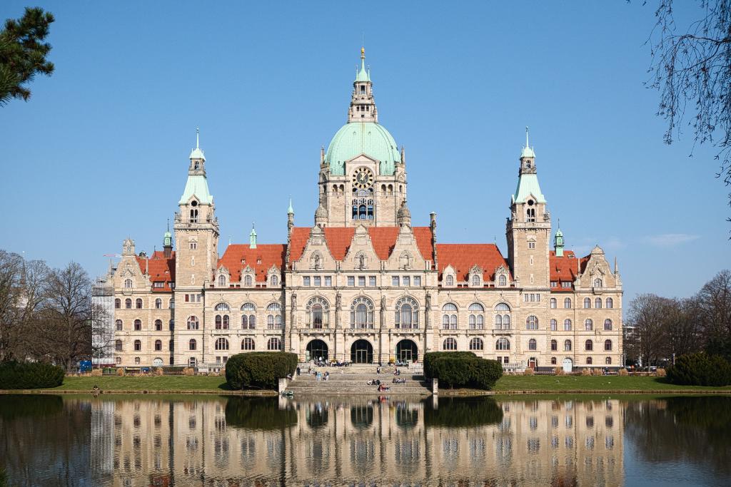 Das Bild zeigt das Rathaus von Hannover an einem sonnigen Tag. Das Gebäude spiegelt sich nahezu perfekt in einem davor liegenden Teich wider. Vor dem Rathaus sitzen einige Personen auf den Treppenstufen. Der Himmel ist klar und blau. Die Architektur des Rathauses ist im Stil der Neogotik gehalten, mit turmartigen Aufbauten und einem auffälligen, kupferfarbenen Dach.
