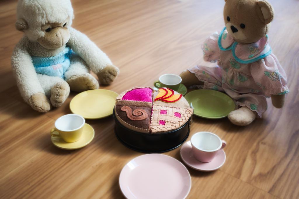 Drei Teller und drei Tassen mit Untertassen in grün, rosa und gelb auf einem Boden in Holzoptik rund um einen Kuchen in einer kleinen Springform angerichtet. Links oben neben dem gelben Teller sitzt ein kleiner weißer Kuscheltieraffe in einer Windel. Rechts oben neben dem grünen Teller sitzt ein Teddybär in einem bunten, antik anmutenden Kleid. Beide gucken auf den Kuchen. Die Kamera ist schräg über dem rosa Teller und blickt ebenfalls zum Kuchen.