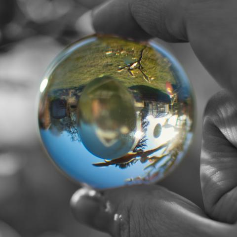 Ein quadratisches Foto einer Glaskugel, die von zwei Fingern oben und unten vor die Kamera gehalten wird. Durch die Glaskugel ist invertiert der Hintergrund erkennbar: Ein Osterei sowie Äste, Himmel und Wiese. Nur die Glaskugel und der Inhalt ist in Farbe, der Rest in Schwarzweiß.