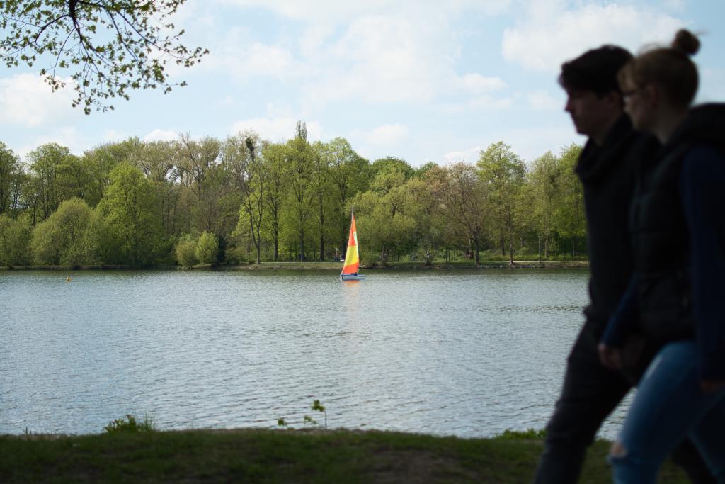 Ein ruhiges Bild eines Sees mit Bäumen am Ufer und zwei Personen, die am Ufer entlang spazieren. Die Bildkomposition ist einfach und konzentriert sich auf die Landschaft. Im Vordergrund ist ein Abschnitt des Ufers mit Gras und Bäumen zu sehen. Im Mittelgrund erstreckt sich ein ruhiger See, auf dem ein kleines Segelboot zu sehen ist. Im Hintergrund erheben sich die Bäume, und der Himmel ist leicht bewölkt, was eine natürliche, unaufgeregte Atmosphäre schafft. Die beiden Personen, die im Vordergrund entlang spazieren, sind leicht verschwommen bzw. unscharf. Die Perspektive ist von einer eher niedrigen Position aus aufgenommen.