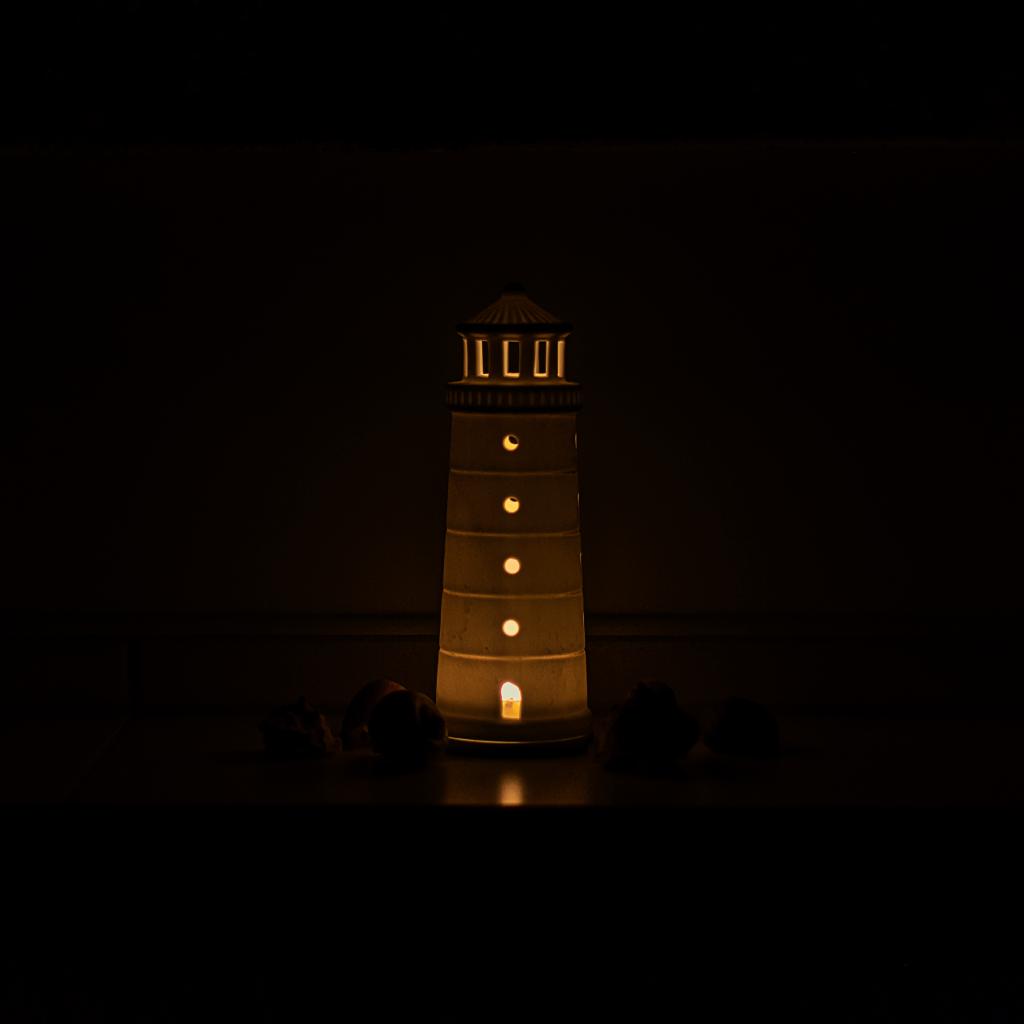 Ein von innen durch eine Kerze erleuchteter, weißer Leuchtturm steht inmitten einer dunklen Umgebung auf einer das Licht leicht spiegelnden Oberfläche. Es sind vor dem Leuchtturm kleine Gegenstände erahnbar wie Steine oder Muscheln. Die Komposition des Bildes ist einfach und fokussiert auf den Leuchtturm. Der Leuchtturm nimmt den größten Teil des Bildes ein und ist der einzige sichtbare Gegenstand.