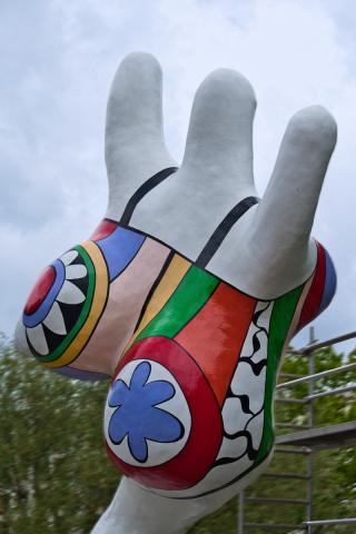 Eine farbenfrohe, abstrakte Skulptur in Form einer Frau mit deutlichen Rundungen, mit bunten Mustern verziert. Die Muster über dem Körper lassen einen an einen Badeanzug denken. Die Skulptur ist von schräg hinten und von unten fotografiert. Es sieht so aus, als würde die Skulptur begeistert und freudig ins Wasser rennen oder hüpfen. Die Skulptur ist als Hauptmotiv präsent und nimmt den Großteil des Bildes ein. Sie ist mit bunten Formen und Farben bedeckt, die in verschiedenen Größen und Mustern angeordnet sind. Die Formen erinnern an geometrische Muster, Kreise, Streifen und Blumen. Die Farben sind leuchtend und kontrastierend, wie Rot, Blau, Grün, Gelb und Weiß. Die Umgebung ist unscharf und die Beleuchtung ist natürlich und scheint von oben zu kommen. Die Hintergrunddetails sind nur vage erkennbar, und sie geben die Atmosphäre von außen in einem Park wieder.