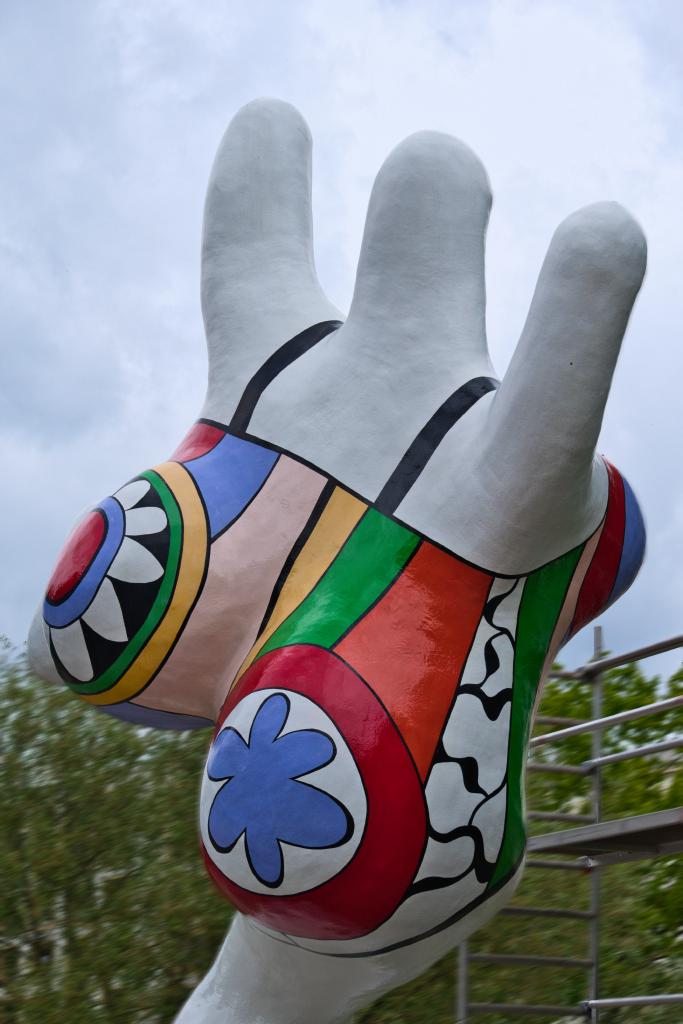 Eine farbenfrohe, abstrakte Skulptur in Form einer Frau mit deutlichen Rundungen, mit bunten Mustern verziert. Die Muster über dem Körper lassen einen an einen Badeanzug denken. Die Skulptur ist von schräg hinten und von unten fotografiert. Es sieht so aus, als würde die Skulptur begeistert und freudig ins Wasser rennen oder hüpfen. Die Skulptur ist als Hauptmotiv präsent und nimmt den Großteil des Bildes ein. Sie ist mit bunten Formen und Farben bedeckt, die in verschiedenen Größen und Mustern angeordnet sind. Die Formen erinnern an geometrische Muster, Kreise, Streifen und Blumen. Die Farben sind leuchtend und kontrastierend, wie Rot, Blau, Grün, Gelb und Weiß. Die Umgebung ist unscharf und die Beleuchtung ist natürlich und scheint von oben zu kommen. Die Hintergrunddetails sind nur vage erkennbar, und sie geben die Atmosphäre von außen in einem Park wieder.