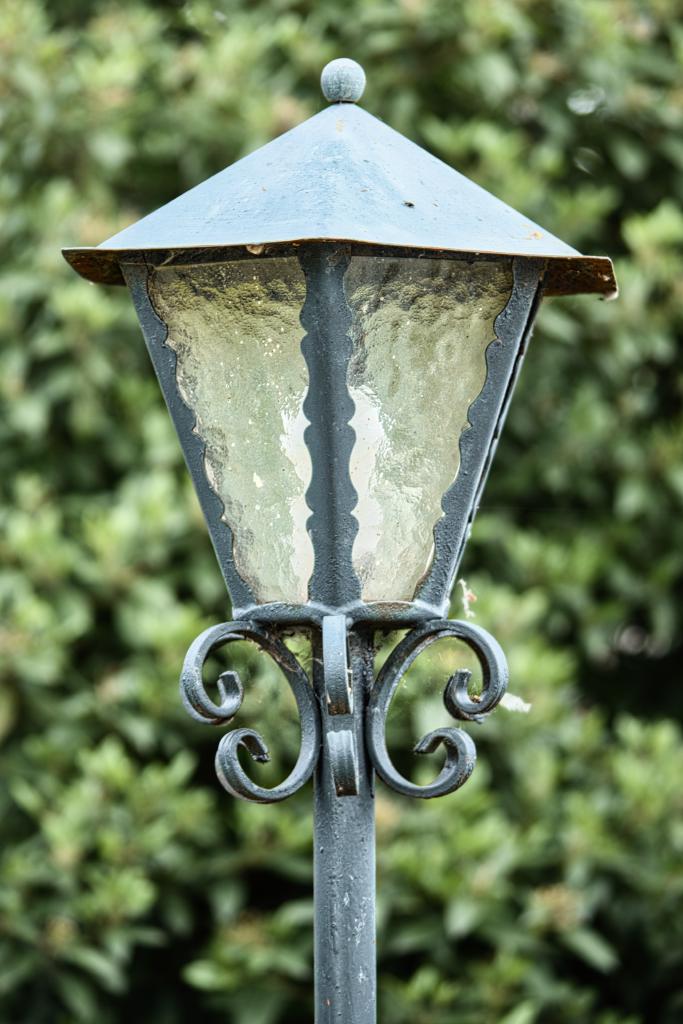 Eine verzierte, quadratische Straßenlaterne mutmaßlich aus Eisen steht vor einem verschwommenen Hintergrund von Bäumen. Die Komposition ist einfach und fokussiert auf die Laterne. Die Laterne nimmt den größten Teil des Bildes ein. Der Hintergrund ist unscharf und stellt einen natürlichen Rahmen für die Laterne dar. Die Positionierung der Elemente ist einfach, da sich die Laterne genau in der Mitte des Bildes befindet. Die Laterne selbst zeigt schmiedeeiserne Verzierungen an der Basis. Die vier Gläser der Laterne sind milchig und undurchsichtig; man sieht nur zwei davon, da die Laterne von der Seite aufgenommen wurde. Die Oberflächenstruktur der Laterne weist Abnutzungs- und Alterungserscheinungen auf, die ihr ein antikes Aussehen verleihen.