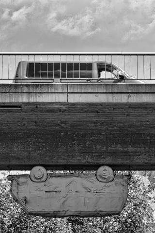 Ein Schwarz-Weiß-Foto zeigt eine Skulptur eines Kleinbus, die unter einer sehr massiven Beton-Brücke hängt. Oben auf der Brücke fährt ebenfalls ein Kleinbus in die entgegengesetzte Richtung. Davor sieht man ein Brückengeländer. Im Hintergrund ist nichts erkennbar außer Himmel und Grünpflanzen. Die Bildkomposition ist minimalistisch und konzentriert sich auf das Auto, das unter der Brücke hängt. Der Fokus liegt auf der ungewöhnlichen Perspektive und der geometrischen Form des Bildes. Das Auto ist in der Mitte des Fotos positioniert und hängt scheinbar in der Luft, was eine unerwartete und interessante Szene erzeugt.