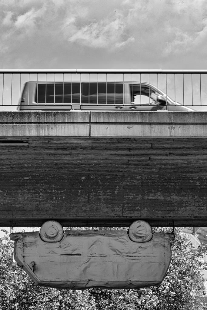 Ein Schwarz-Weiß-Foto zeigt eine Skulptur eines Kleinbus, die unter einer sehr massiven Beton-Brücke hängt. Oben auf der Brücke fährt ebenfalls ein Kleinbus in die entgegengesetzte Richtung. Davor sieht man ein Brückengeländer. Im Hintergrund ist nichts erkennbar außer Himmel und Grünpflanzen. Die Bildkomposition ist minimalistisch und konzentriert sich auf das Auto, das unter der Brücke hängt. Der Fokus liegt auf der ungewöhnlichen Perspektive und der geometrischen Form des Bildes. Das Auto ist in der Mitte des Fotos positioniert und hängt scheinbar in der Luft, was eine unerwartete und interessante Szene erzeugt.