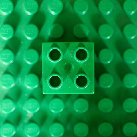 Die Szene zeigt einen einzelnen, quadratischen grünen LEGO-Stein, der auf einer größeren LEGO-Platte derselben Farbe steckt. Der Stein ist in der Mitte des Bildes positioniert. Die Anordnung erzeugt ein geometrisches Muster. Der Stein und die Platte zeigen eine gleichmäßige Färbung und Oberflächenstruktur. Stein und Platte haben deutlich sichtbare runde Löcher auf jeder Seite, die für die Verbindung mit anderen LEGO-Steinen gedacht sind.