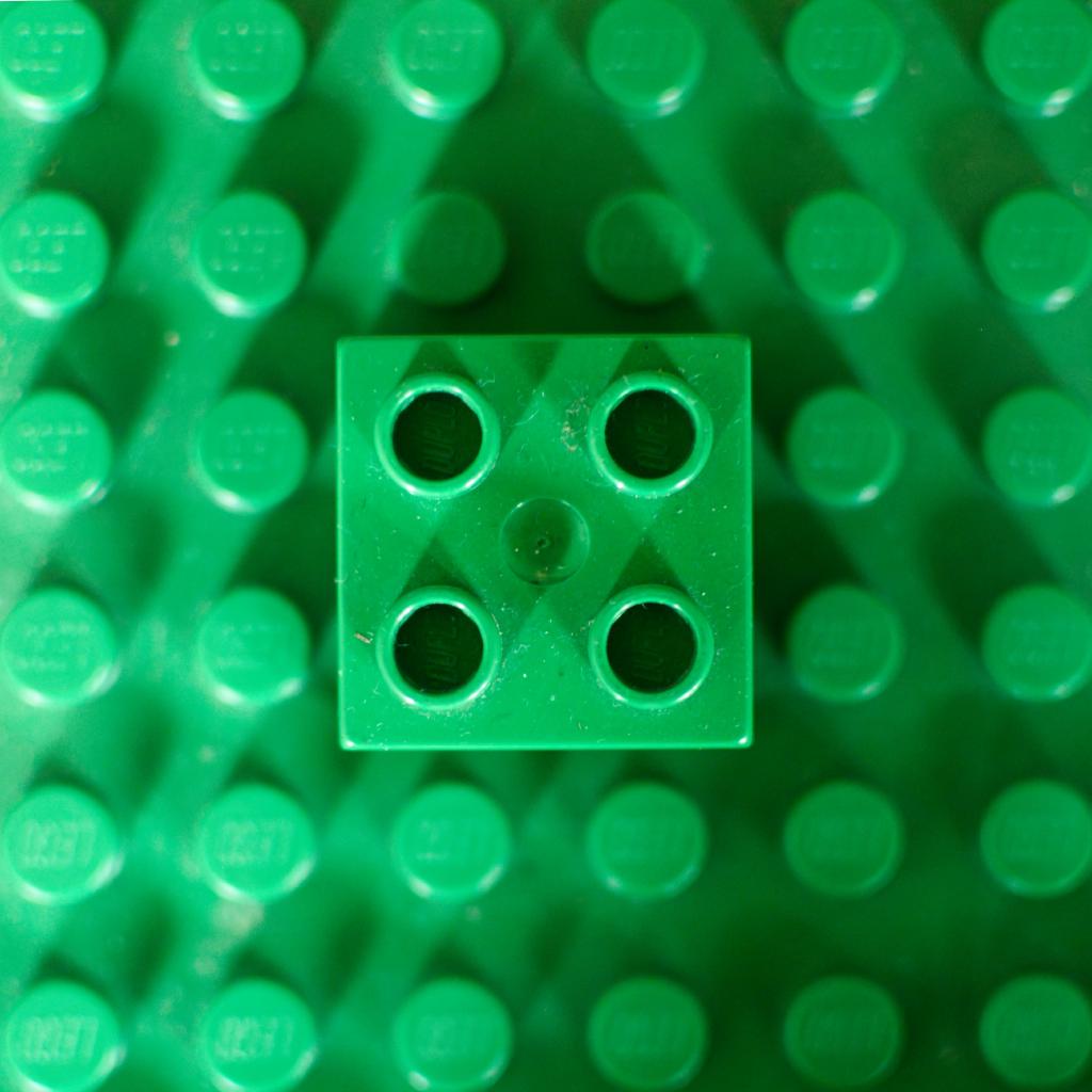 Die Szene zeigt einen einzelnen, quadratischen grünen LEGO-Stein, der auf einer größeren LEGO-Platte derselben Farbe steckt. Der Stein ist in der Mitte des Bildes positioniert. Die Anordnung erzeugt ein geometrisches Muster. Der Stein und die Platte zeigen eine gleichmäßige Färbung und Oberflächenstruktur. Stein und Platte haben deutlich sichtbare runde Löcher auf jeder Seite, die für die Verbindung mit anderen LEGO-Steinen gedacht sind.