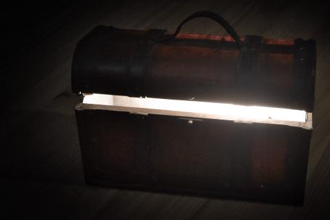 Ein braunes, altes Truhen-Objekt steht mit leicht offenem Deckel auf einer Art Holzfußboden, wobei ein heller Lichtstrahl aus der Truhe heraus scheint. Die Komposition zeigt eine Truhe, die in der Mitte des Bildes positioniert ist. Die Truhe ist von einem dunklen Hintergrund bzw. einer Vignette umgeben, was den Kontrast zum hellen Lichtinneren verstärkt. Die Truhe hat eine klassische Truhenform mit einem Deckel und Griffen, die das Objekt mit einem traditionellen Aussehen ausstatten. Ein Licht scheint von innen heraus zu leuchten.