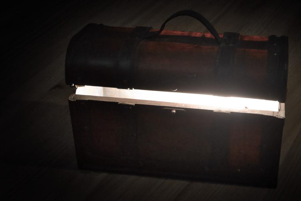 Ein braunes, altes Truhen-Objekt steht mit leicht offenem Deckel auf einer Art Holzfußboden, wobei ein heller Lichtstrahl aus der Truhe heraus scheint. Die Komposition zeigt eine Truhe, die in der Mitte des Bildes positioniert ist. Die Truhe ist von einem dunklen Hintergrund bzw. einer Vignette umgeben, was den Kontrast zum hellen Lichtinneren verstärkt. Die Truhe hat eine klassische Truhenform mit einem Deckel und Griffen, die das Objekt mit einem traditionellen Aussehen ausstatten. Ein Licht scheint von innen heraus zu leuchten.