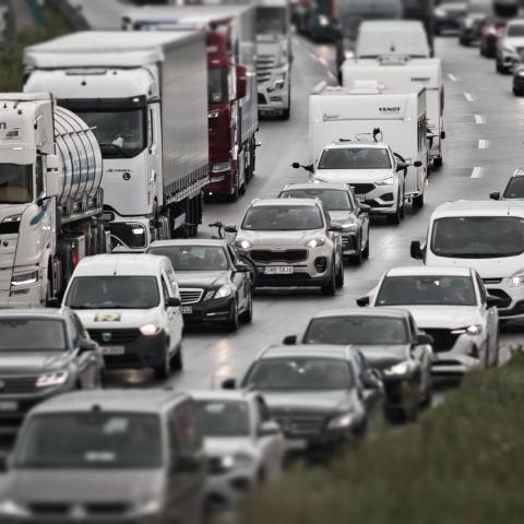 Ein Bild zeigt einen Stau auf einer Autobahn. Die Bildkomposition zeigt eine Vielzahl von Fahrzeugen, darunter Autos und LKWs, die sich auf einer Autobahn in einem Stau befinden. Die Fahrzeuge sind dicht aneinandergereiht, und die Straße ist mit Regen bedeckt. Die Perspektive ist von oben, und der Blick geht von vorne nach hinten über die Autobahn. Die Beleuchtung ist diffus, da es regnet und keine starke Sonnenlichtquelle vorhanden ist. Die Atmosphäre ist ruhig und die Stimmung ist ein wenig nachdenklich.