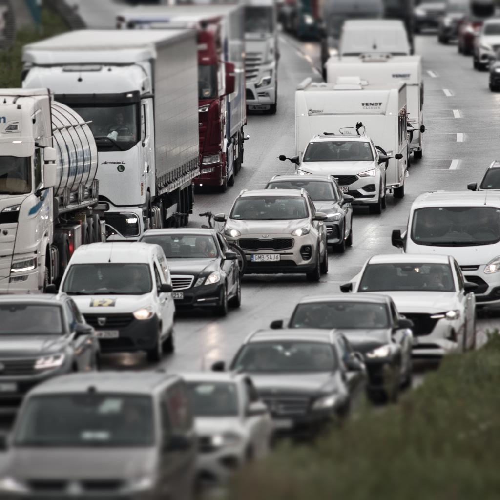 Ein Bild zeigt einen Stau auf einer Autobahn. Die Bildkomposition zeigt eine Vielzahl von Fahrzeugen, darunter Autos und LKWs, die sich auf einer Autobahn in einem Stau befinden. Die Fahrzeuge sind dicht aneinandergereiht, und die Straße ist mit Regen bedeckt. Die Perspektive ist von oben, und der Blick geht von vorne nach hinten über die Autobahn. Die Beleuchtung ist diffus, da es regnet und keine starke Sonnenlichtquelle vorhanden ist. Die Atmosphäre ist ruhig und die Stimmung ist ein wenig nachdenklich.