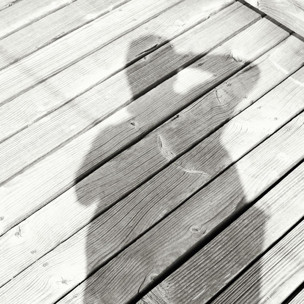 Ein Schwarzweiß-Foto zeigt den Schatten einer Person auf einem Holzboden oder einer Holzterrasse. Die Holzplanken verlaufen in einer Reihe und zeigen eine lineare Musterung. Die Schattenform ist unregelmäßig und gibt die Silhouette des Körpers an. Die Beleuchtung und das Licht scheinen von oben oder seitlich zu kommen.