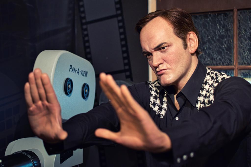 Eine detaillierte Wachsfiguren-Nachbildung von Quentin Tarantino, der eine Geste mit beiden Händen macht, wobei er sie vor seinen Augen in Form einer imaginären Leinwand formt, um die Perspektive einer Szene abzuschätzen. Die Komposition des Bildes ist auf ihn konzentriert. Eine alte Filmkamera nimmt einen Teil des Hintergrunds ein. Das Aussehen von Tarantino, inklusive seiner Gesichtszüge und Kleidung, ist in den Details realistisch dargestellt. Sein Ausdruck suggeriert Konzentration.