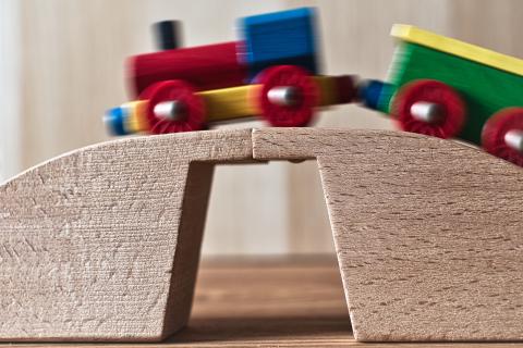 Ein detaillierter Blick auf ein hölzernes Spielzeug-Viadukt, über das eine bunte Lok mit einem Waggen fährt. Der Vordergrund konzentriert sich auf die Struktur des Holzviadukts, während der Hintergrund die verschwommenen Züge zeigt, die über das Viadukt fahren. Die Züge sind in Bewegung, was durch die unscharfe Darstellung der Züge im Bild vermittelt wird. Die Positionierung der Züge im Bild konzentriert sich auf die Bewegung und Dynamik der Szene. Der Hintergrund ist hell und neutral. Die Beleuchtung ist einfach und dient nur der Darstellung der Züge und des Viadukts.