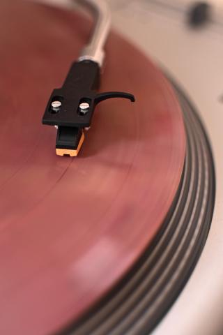 Das Foto zeigt eine Nahaufnahme eines Plattenspielers, wobei der Tonabnehmer auf einer sich drehenden Schallplatte in einem sanften, hellroten Ton liegt. Die Komposition konzentriert sich auf das Detail des Plattenspielers, wobei der Tonabnehmer im Mittelpunkt steht und über der sich drehenden Schallplatte schwebt. Die Platte wird im Detail dargestellt, mit sichtbaren Rillen und der reflektierenden Oberfläche. Der Winkel der Aufnahme ist leicht von oben, wodurch die Bewegung und die mechanische Komplexität des Geräts hervorgehoben werden.