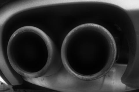 Das Schwarzweißfoto zeigt zwei Auspuffrohre eines Autos, die von unten aus einem leicht schrägen Winkel aufgenommen wurden, wodurch die zylindrischen Formen betont werden. Die Komposition ist einfach und auf die Geometrie der Auspuffrohre ausgerichtet, wobei die beiden Rohre im Zentrum des Bildes dominieren.