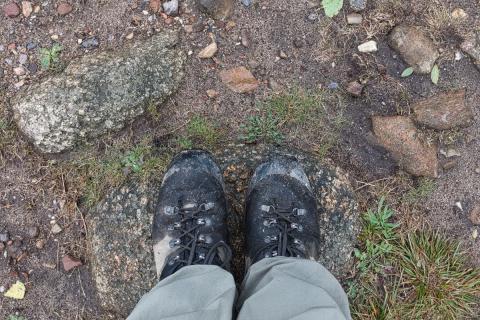 Die Nahaufnahme zeigt die Füße einer Person, die auf einem unebenen Untergrund aus Steinen, Erde und Gras steht und robuste, verschmutzte Wanderschuhe trägt. Die Szene ist aus der Ich-Perspektive fotografiert und zeigt die Füße der Person von oben, wobei der Blick nach unten auf den Boden gerichtet ist. Die Komposition ist relativ symmetrisch, mit den Schuhspitzen im Zentrum und den hellbraunen Hosenbeinen, die leicht nach unten zusammenlaufen.