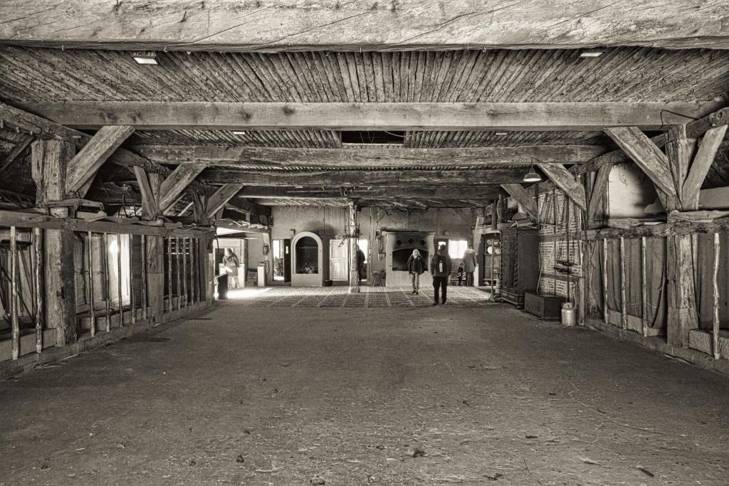 Das Schwarz-Weiß-Foto mit Sepia-Tönung zeigt den Innenraum eines historischen Gebäudes, vermutlich ein altes Landhaus, mit hölzernen Strukturen, die eine stimmungsvolle Atmosphäre erzeugen. Im vorderen Bereich der Kamera sind links und rechts Stallungen sichtbar und in der Mitte so viel Platz, dass man von einem riesigen Saal sprechen kann. Im Hintergrund am anderen Ende des Raums lassen sich ein alter Ofen sowie Türen in den Wohnbereich des alten Landhauses erkennen. Die Komposition ist symmetrisch aufgebaut, wobei die zentralen Elemente auf die Mitte ausgerichtet sind und das Auge des Betrachters in die Tiefe des Raumes führen. Das Hauptaugenmerk liegt auf dem ausladenden Dachgebälk und den Stützbalken, die das Gebäude tragen. Mehrere Personen sind in der Ferne zu erkennen, die sich scheinbar im Raum bewegen oder ihn betreten.