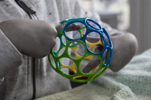 Ein Nahaufnahmefoto zeigt die Hände eines Kindes, das einen mehrfarbigen Gitterball hält, vor einem unscharfen, grauweißen Hintergrund, der die lebendigen Farben des Balls hervorhebt. Die Komposition ist nah und konzentriert sich auf die Hände des Kindes, das den bunten Spielball hält, die Hände sind im Bildzentrum positioniert, wodurch die Aufmerksamkeit auf das Objekt gelenkt wird. Die unscharfe, monochromatische Umgebung dient als Hintergrund und lenkt die Aufmerksamkeit auf die lebendigen Farben des Spielzeugs. Der Bildausschnitt ist eng, der Fokus liegt auf dem Spielball und den Händen, wodurch ein Gefühl der Intimität erzeugt wird. Die Hauptelemente sind die Hände und der Gitterball, das Kind ist nicht vollständig zu sehen, nur die Hände, die den farbenfrohen Ball halten. Der Gitterball hat verschiedene leuchtende Farben, grün, gelb und blau, die in einem Raster angeordnet sind. Die Hände scheinen klein zu sein, was darauf schließen lässt, dass das Kind jung ist.