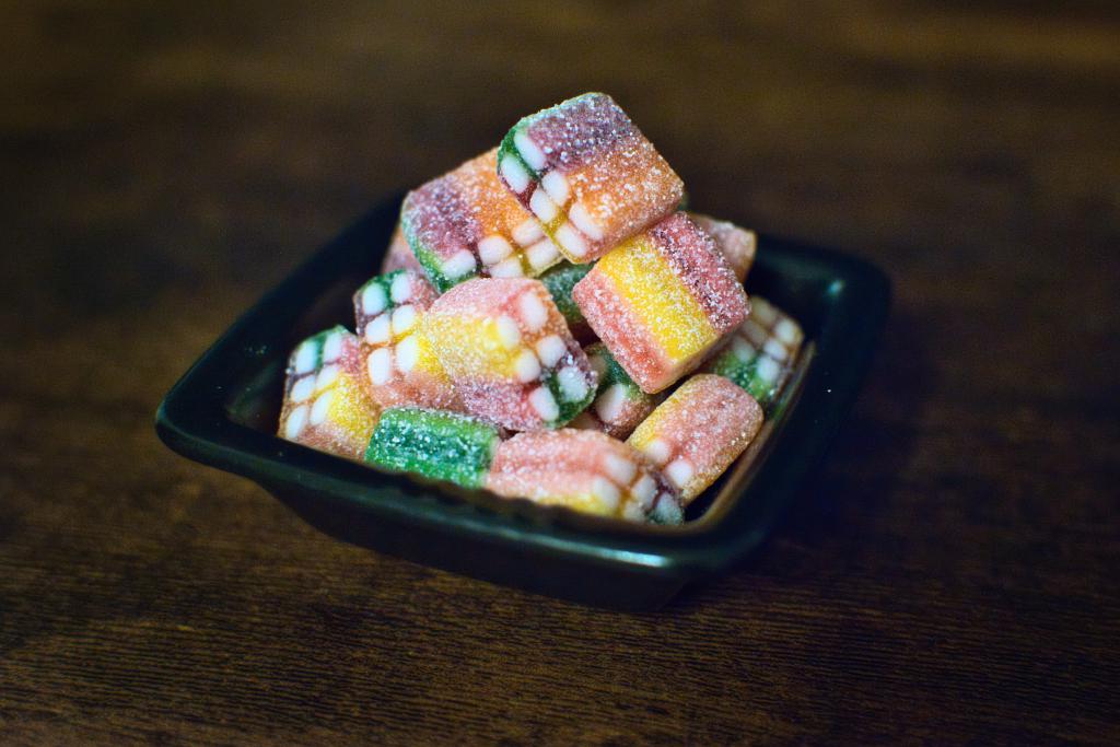 Ein Stillleben zeigt eine kleine schwarze Schale, gefüllt mit farbenfrohen, zuckerbedeckten Fruchtgummis, die auf einem dunklen Holzhintergrund steht. Die Komposition ist schlicht und fokussiert, wobei die Schale mit den Süßigkeiten den größten Teil des Bildes einnimmt. Die Schale ist leicht von oben gesehen, was die Form der quadratischen Fruchtgummis und ihre Anordnung in der Schale betont. Die Fruchtgummis sind unordentlich gestapelt, was ein Gefühl von Fülle und Verlockung erzeugt. Der Hintergrund ist ein einfacher, dunkler Holztisch, der die leuchtenden Farben der Süßigkeiten hervorhebt. Die Beleuchtung scheint natürlich zu sein, wobei das Licht von oben kommt und leichte Schatten auf den Fruchtgummis und der Schale wirft, die Tiefe erzeugen. Die Fruchtgummis sind quadratisch geformt und in verschiedenen Farben gestaltet, darunter Grün, Gelb, Orange und Rosa. Jedes Fruchtgummi scheint mit Zucker bedeckt zu sein, was ihm eine glänzende Textur verleiht.