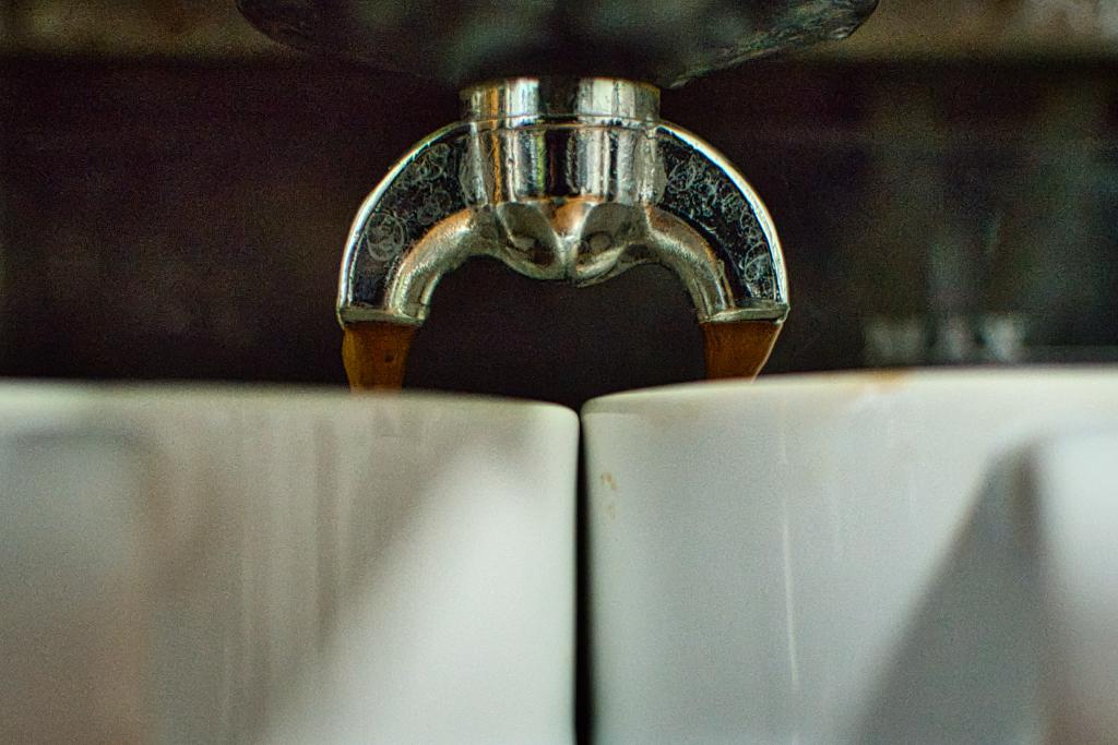 Das Bild zeigt eine Nahaufnahme, in der Kaffee aus einem Espressomaschinen-Auslauf in zwei weiße Tassen fließt, die sich darunter befinden. Die Szene ist eng gefasst und konzentriert sich auf das Zentrum der Espressomaschine. Der metallische Auslauf teilt das Bild horizontal in zwei Hälften und leitet das fließende dunkle Getränk in die darunter stehenden Tassen. Die Komposition ist symmetrisch, wobei die beiden Tassen und der Auslauf in einer ausgewogenen Anordnung positioniert sind. Der Hintergrund ist unscharf, was den Fokus auf die Hauptobjekte verstärkt und dem Bild Tiefe verleiht. Die Textur des Kaffees ist sichtbar, was das Gefühl von Frische und Wärme erzeugt.