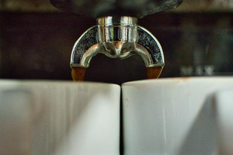 Das Bild zeigt eine Nahaufnahme, in der Kaffee aus einem Espressomaschinen-Auslauf in zwei weiße Tassen fließt, die sich darunter befinden. Die Szene ist eng gefasst und konzentriert sich auf das Zentrum der Espressomaschine. Der metallische Auslauf teilt das Bild horizontal in zwei Hälften und leitet das fließende dunkle Getränk in die darunter stehenden Tassen. Die Komposition ist symmetrisch, wobei die beiden Tassen und der Auslauf in einer ausgewogenen Anordnung positioniert sind. Der Hintergrund ist unscharf, was den Fokus auf die Hauptobjekte verstärkt und dem Bild Tiefe verleiht. Die Textur des Kaffees ist sichtbar, was das Gefühl von Frische und Wärme erzeugt.