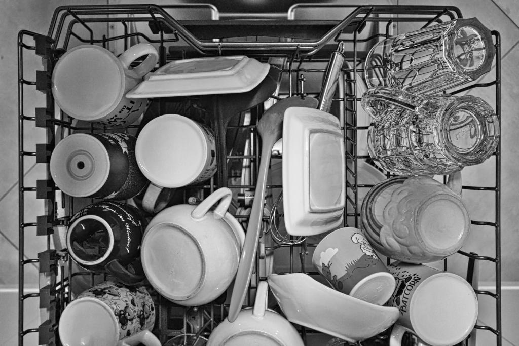 Das Schwarz-Weiß-Foto zeigt eine Draufsicht auf den Innenraum eines Geschirrspülers, der mit einer Vielzahl von Geschirr, wie Tassen, Schalen und Gläsern, gefüllt ist. Die Komposition ist durch die Aufsicht geprägt und zeigt einen dicht gepackten Geschirrspüler mit einer Vielzahl von Objekten, die scheinbar unordentlich, aber funktional angeordnet sind. Die Objekte sind scheinbar willkürlich in den dafür vorgesehenen Halterungen platziert, wobei die Platzierung durch die begrenzten räumlichen Kapazitäten des Geschirrspülers bestimmt wird. Die Hauptobjekte sind eine Vielzahl von Geschirrteilen, darunter Tassen in verschiedenen Größen und Designs, flache Teller, kleine Schalen, Gläser und Servierlöffel. Einige Tassen weisen dekorative Muster auf, während andere schlicht einfarbig sind. Die Gläser haben eine transparente Struktur, die die Reflexion von Licht in den Spaltrichtungen hervorhebt. Das Geschirr scheint sauber, und der Waschvorgang scheint gerade abgeschlossen.