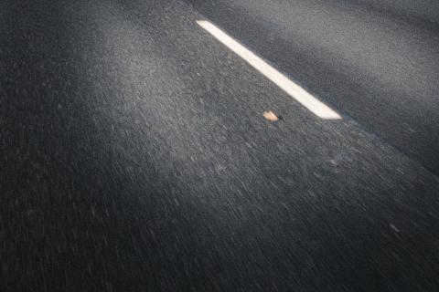 Die Aufnahme zeigt eine Nahaufnahme einer asphaltierten Straße mit einer weißen Mittellinie, die leicht von oben links nach unten rechts verläuft, sowie einen kleinen Gegenstand, mutmaßlich ein Blatt. Die Bildkomposition fokussiert sich auf die Textur der Straße und die diagonale Anordnung der Mittellinie. Der Blickwinkel ist leicht von oben, was dem Betrachter das Gefühl vermittelt, sich auf der Straße fortzubewegen, was durch die Bewegungsunschärfe verstärkt wird. Die Mittellinie dient als visuelle Führung, die den Blick des Betrachters durch das Bild lenkt. Die Platzierung des kleinen Objekts neben der Linie fügt der ansonsten abstrakten Szene einen Hauch von Realismus hinzu. Der Schwerpunkt des Bildes liegt nicht auf bestimmten Objekten, sondern auf der Beschaffenheit der Straße und der weißen Linie.