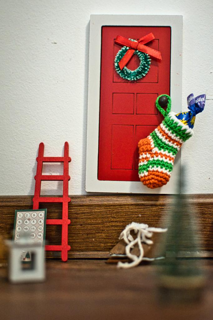Dieses Foto zeigt eine miniaturhafte Szene mit weihnachtlichen Dekorationen, darunter ein gestrickter Strumpf, eine rote Wichtel-Tür mit Kranz an einer Wand hängend und eine kleine, rote Leiter. Die Komposition ist vertikal angeordnet, wobei die Hauptobjekte wie die rote Tür, der Strumpf und die Leiter in einer leicht asymmetrischen, aber ausgewogenen Weise angeordnet sind. Die Szene wirkt wie ein Miniatur-Set oder eine Installation. Der Blick des Betrachters wird durch die rote Tür sowie die Leiter angezogen, während andere Elemente wie ein verschwommen im Vordergrund stehender Weihnachtsbaum und eine kleine Kalenderkarte eine unterstützende Rolle spielen. Die Anordnung schafft eine gemütliche und spielerische Atmosphäre.