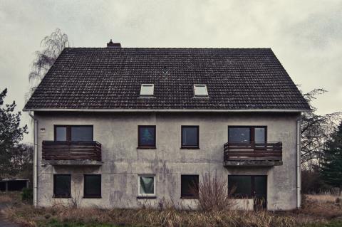 Das Bild zeigt ein zweistöckiges, grau-weißes Haus in einem verlassenen Zustand, das unter einem trüben Himmel steht und eine melancholische Atmosphäre ausstrahlt. Die Komposition ist symmetrisch, wobei das Haus im Mittelpunkt steht, umgeben von einem kargen Vordergrund mit trockenem Gras und Gestrüpp, das zu den Seiten hin verschwimmt. Das Gebäude ist zentriert, mit einem Satteldach und zwei Balkonen an den oberen Fenstern. Die Bildausrichtung ist frontal, was die Struktur direkt präsentiert. Das Haus weist sichtbare Zeichen des Alterns auf, wie beispielsweise durch Verwitterung und Abnutzung. Die Fenster sind dunkel und einige spiegeln das trübe Wetter wider, während in einem Fenster ein weihnachtsähnliches Bild zu sehen ist, das einen Kontrast zur allgemeinen Stimmung erzeugt. Die Balkone sind mit einfachen Holzgeländern ausgestattet und die Dachziegel sind dunkel und möglicherweise mit Flechten bedeckt.