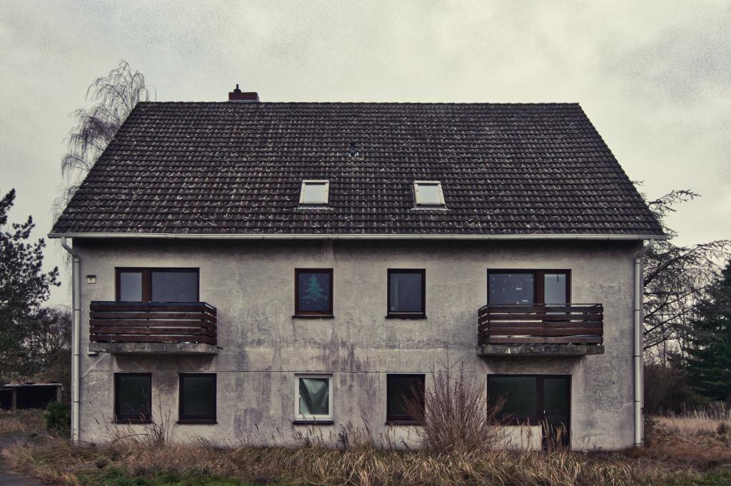 Das Bild zeigt ein zweistöckiges, grau-weißes Haus in einem verlassenen Zustand, das unter einem trüben Himmel steht und eine melancholische Atmosphäre ausstrahlt. Die Komposition ist symmetrisch, wobei das Haus im Mittelpunkt steht, umgeben von einem kargen Vordergrund mit trockenem Gras und Gestrüpp, das zu den Seiten hin verschwimmt. Das Gebäude ist zentriert, mit einem Satteldach und zwei Balkonen an den oberen Fenstern. Die Bildausrichtung ist frontal, was die Struktur direkt präsentiert. Das Haus weist sichtbare Zeichen des Alterns auf, wie beispielsweise durch Verwitterung und Abnutzung. Die Fenster sind dunkel und einige spiegeln das trübe Wetter wider, während in einem Fenster ein weihnachtsähnliches Bild zu sehen ist, das einen Kontrast zur allgemeinen Stimmung erzeugt. Die Balkone sind mit einfachen Holzgeländern ausgestattet und die Dachziegel sind dunkel und möglicherweise mit Flechten bedeckt.