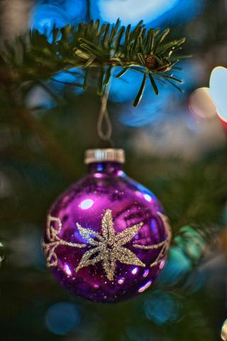 Die Nahaufnahme eines Weihnachtsbaums zeigt eine glänzende, violette Christbaumkugel, die an einem Ast hängt und von farbenfrohen Lichtern umgeben ist, wodurch eine festliche Atmosphäre entsteht. Die Komposition ist vertikal ausgerichtet und präsentiert die violette Christbaumkugel als Hauptobjekt in der Bildmitte, die an einem Zweig eines Weihnachtsbaums befestigt ist. Die Kugel ist leicht nach links gerichtet, während der Zweig von oben ins Bild ragt. Der Hintergrund ist unscharf gehalten und zeigt farbige Lichter, die einen Bokeh-Effekt erzeugen. Die Hauptfigur ist eine runde, glänzende Christbaumkugel in tiefem Violett, die mit einem sternförmigen Muster aus glitzerndem Silber verziert ist. Das Metall der Kugel ist stark reflektierend und spiegelt die Umgebung, wodurch Lichtreflexe und eine sanfte Farbverteilung entstehen. Der grüne Zweig, an dem die Kugel hängt, ist mit Nadeln bedeckt und verleiht dem Bild ein natürliches Element.